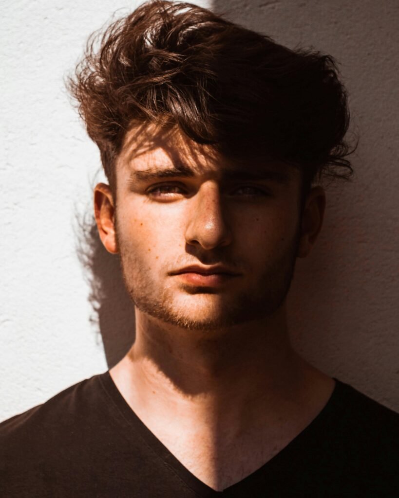 Artistic portrait of a young man in dramatic lighting with half face in shadow, against a textured wall.